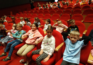 Na zdjęciu dzieci siedzące na czerwonych fotelach w sali teatralnej.